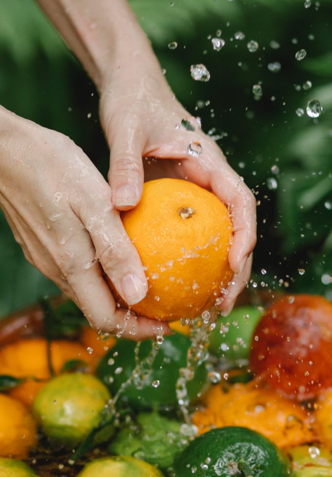 Algumas frutas cítricas estão na bacia com água, em quanto a pessoa está lavando a laranja da variedade bahia.